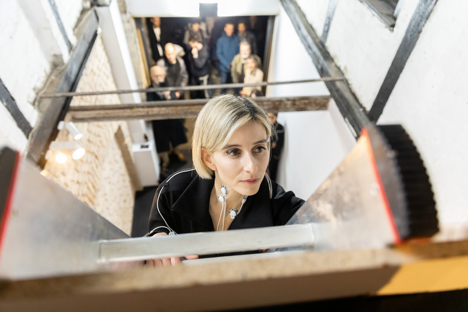 Katz's face low between two angled forms with a glass cylinder resting across them, the audience out of focus above her