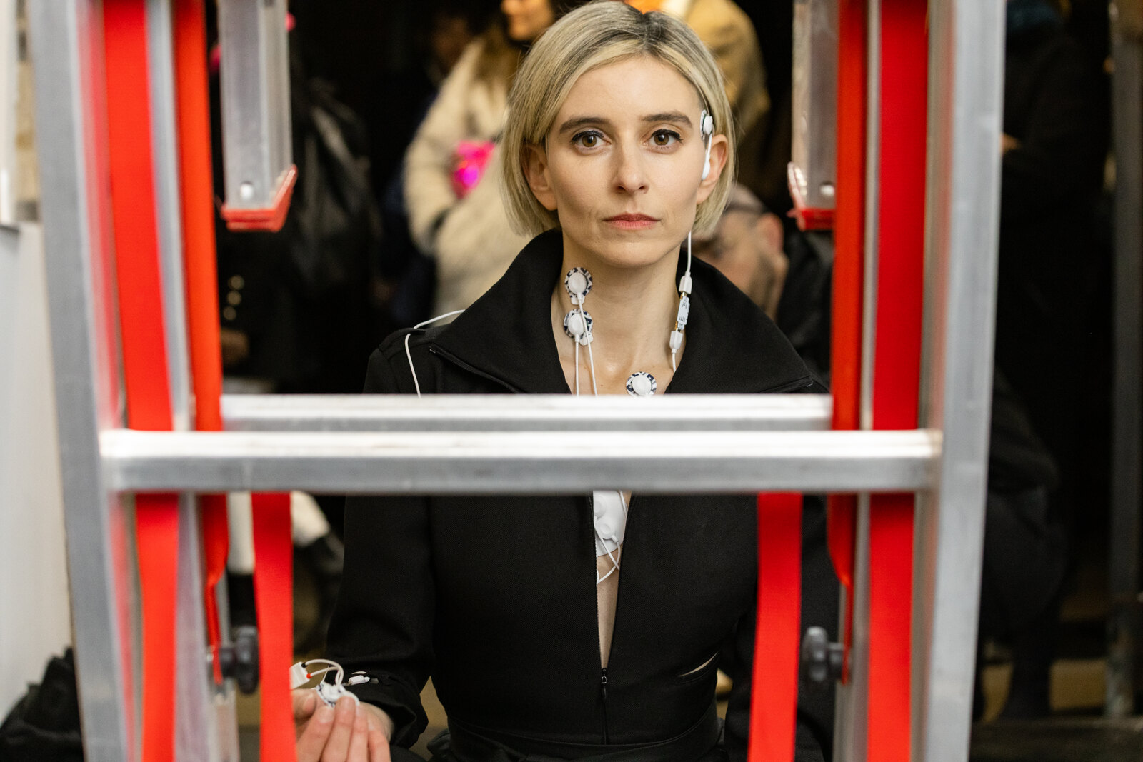 Elana Katz framed inside the red uprights of the ladder, electrodes and leads on her face and neck, looking straight out at the camera
