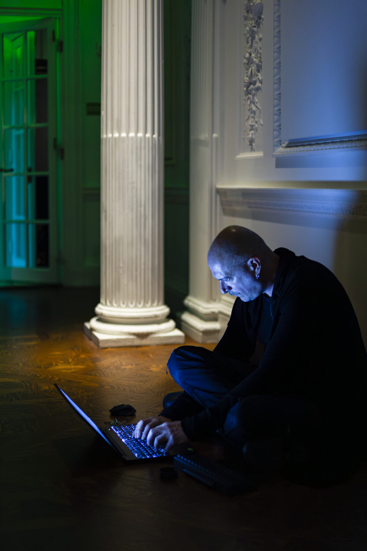 The artist crouched on the parquet beside a column with a laptop open on the floor, green and blue light across the wall behind him