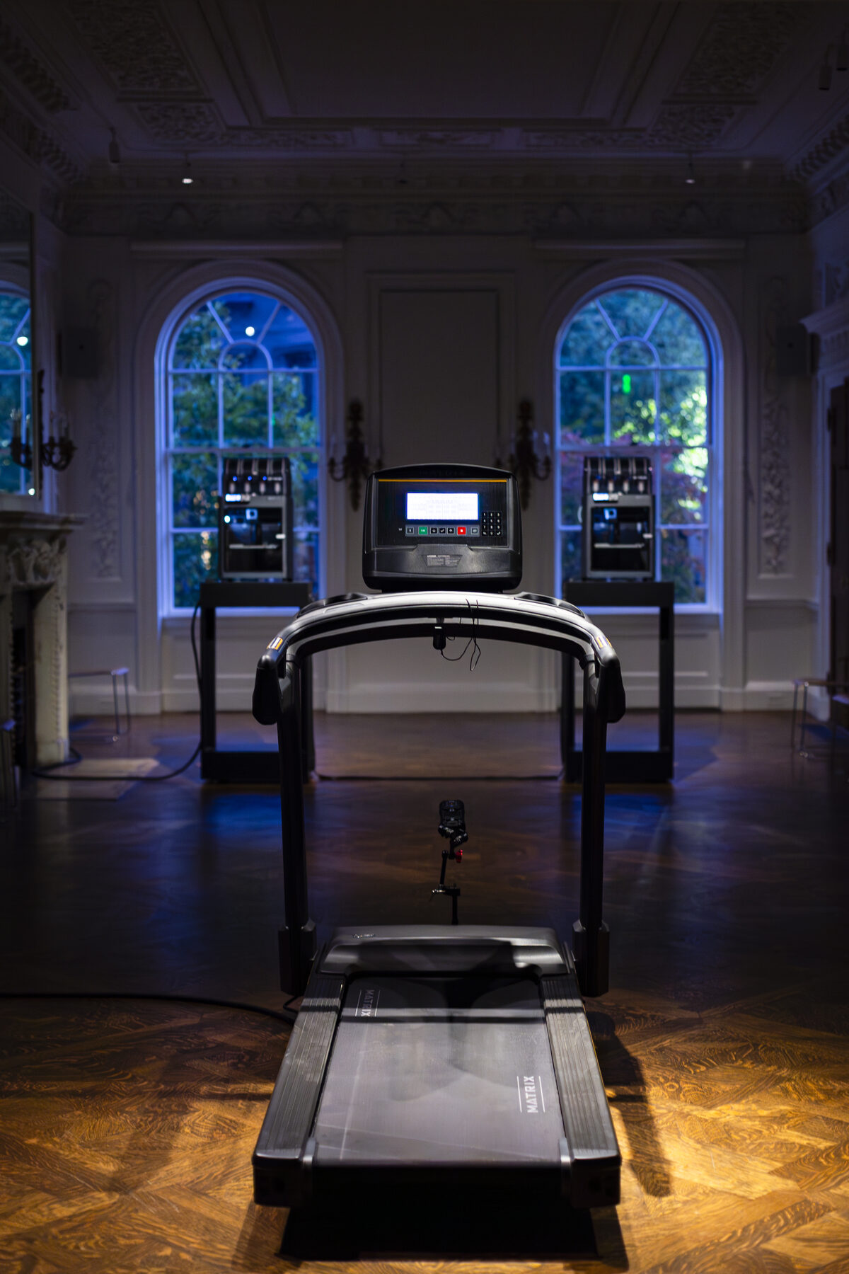 An empty treadmill lit alone on the parquet of a tall panelled hall, the two printers standing between arched windows behind it under blue light