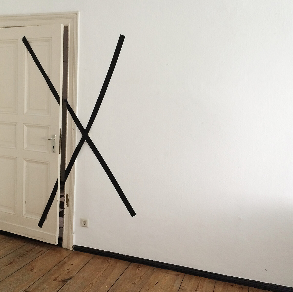 A white room with bare floorboards, an open panelled door, and a large X taped across the doorway in black tape