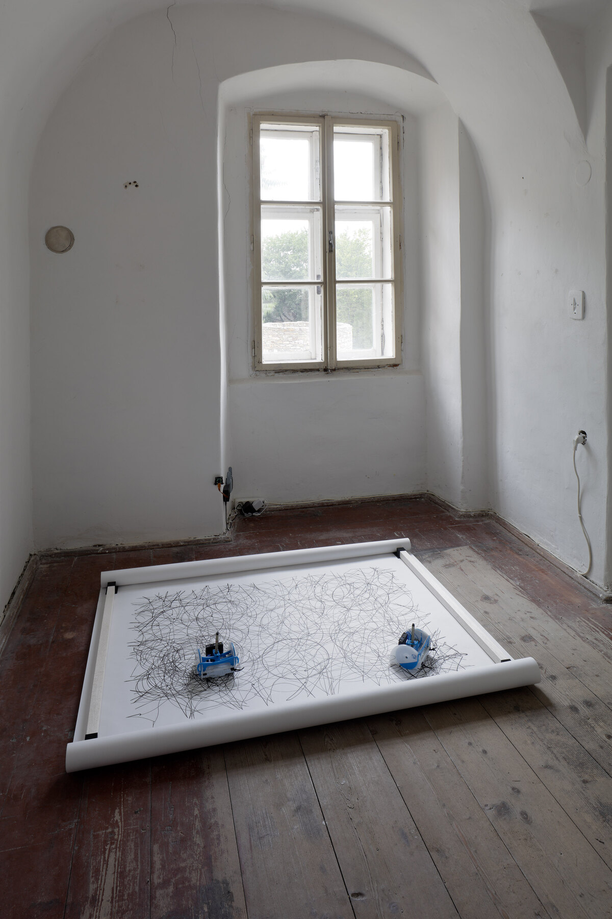A small vaulted whitewashed room with a single window onto trees, the paper band laid on the bare plank floor with both robots at rest on it
