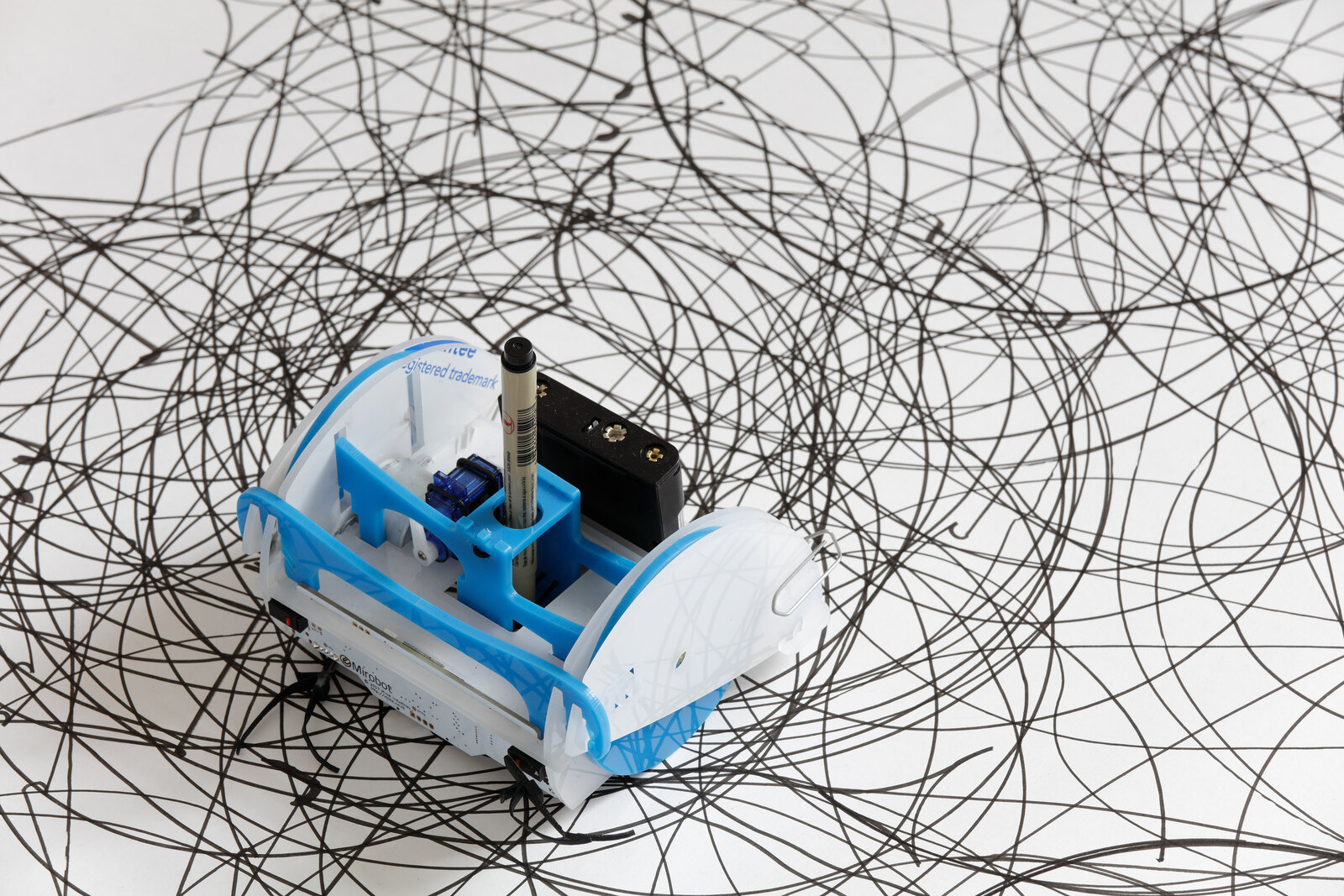 One robot close up, resting on its own accumulated trace, the brush pen upright in its holder and the ink lines falling out of focus behind it