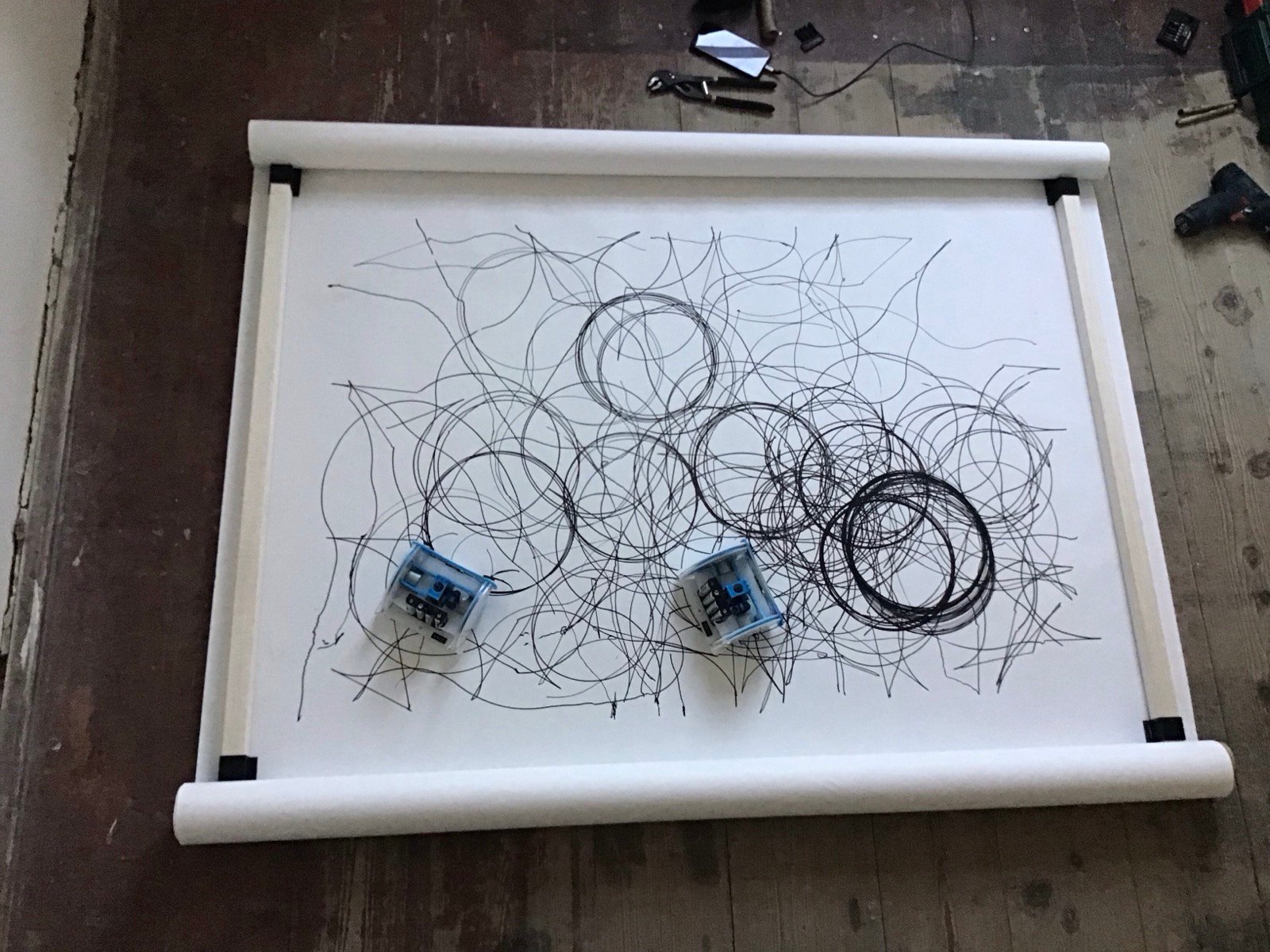 A band marked with a loose scatter of thin circles and, at its centre, two heavy black rings of roughly equal size overlapping in a shared lens; both robots on the paper, pliers and a cordless drill still lying on the floor alongside