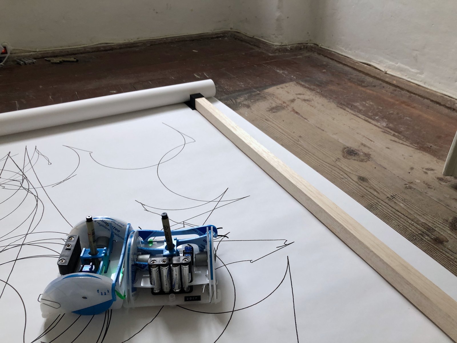 The near corner of the band: the paper roll, the batten clamped to it, a robot at the edge on a field of thin lines, the plank floor beyond