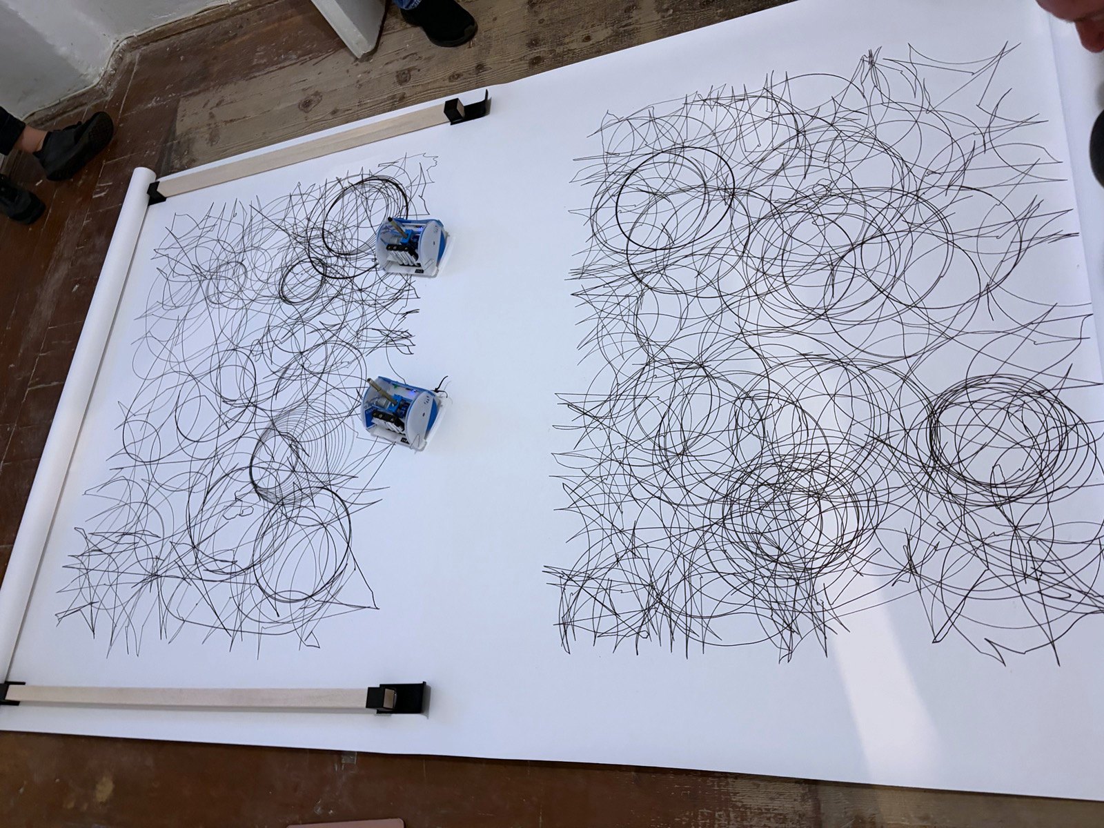 The roll unrolled along the plank floor: two finished drawings side by side, each a dense field of overlapping circles and lines, both robots at rest at the right edge of the left one