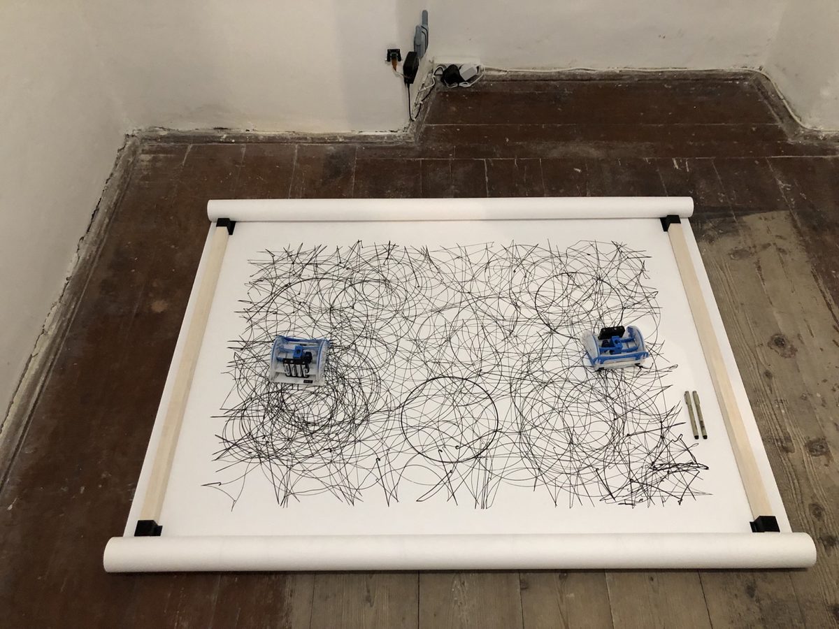 Two drawing robots at rest on a paper roll covered in overlapping ink circles, clamped between wooden battens on a bare plank floor