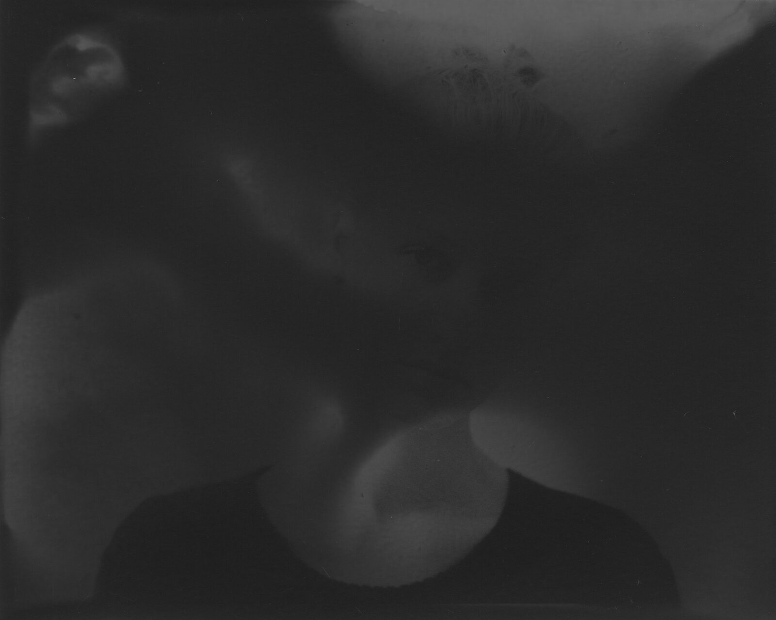 A very dark black-and-white analogue print in which a face surfaces from the darkness to a different degree
