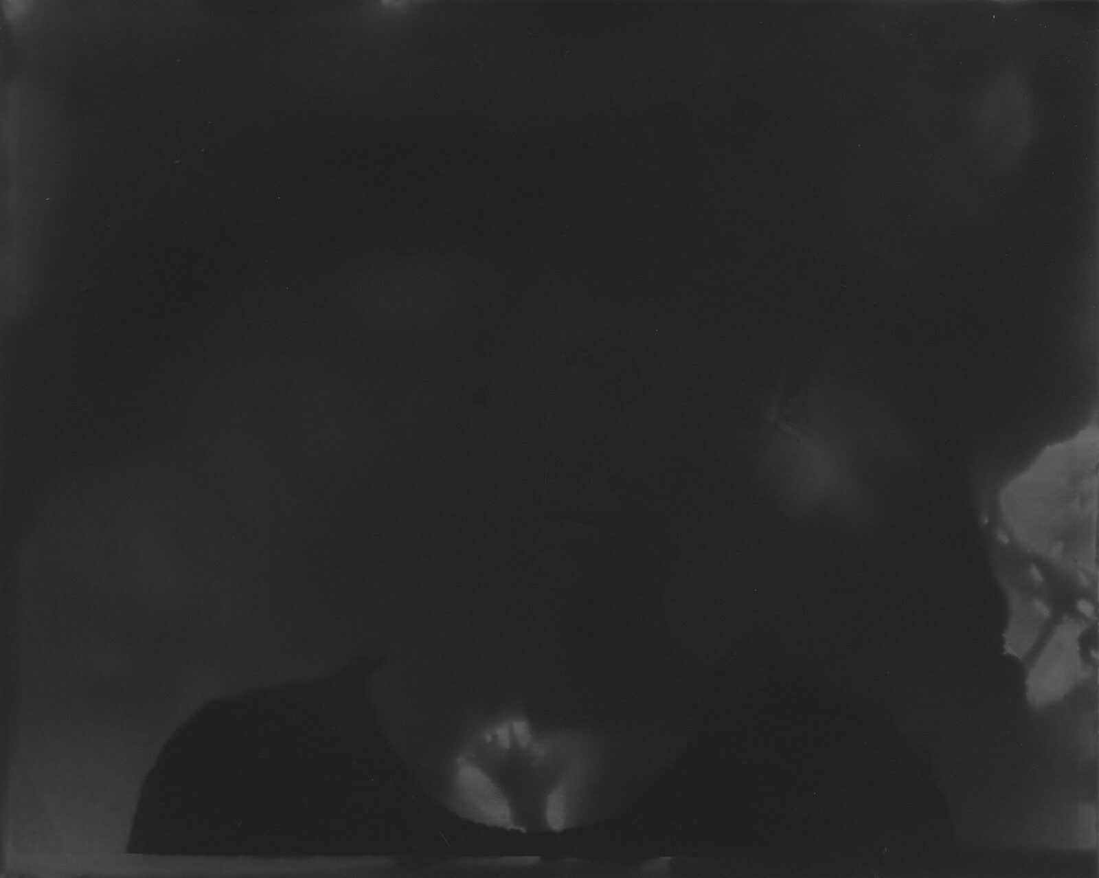 A very dark black-and-white analogue print in which a face surfaces from the darkness to a different degree