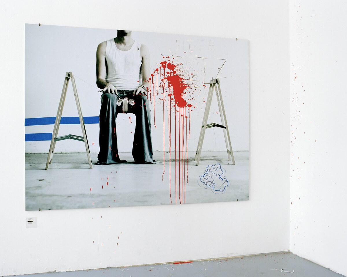 A large mounted photograph of a seated figure between two trestles, struck with red paint that runs off the print onto the gallery wall and floor, a small wall label beside it
