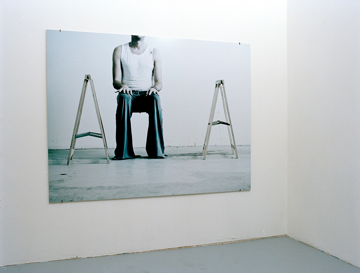 The untouched photograph mounted alone on a white gallery wall: a seated figure in a white tank top between two wooden trestles