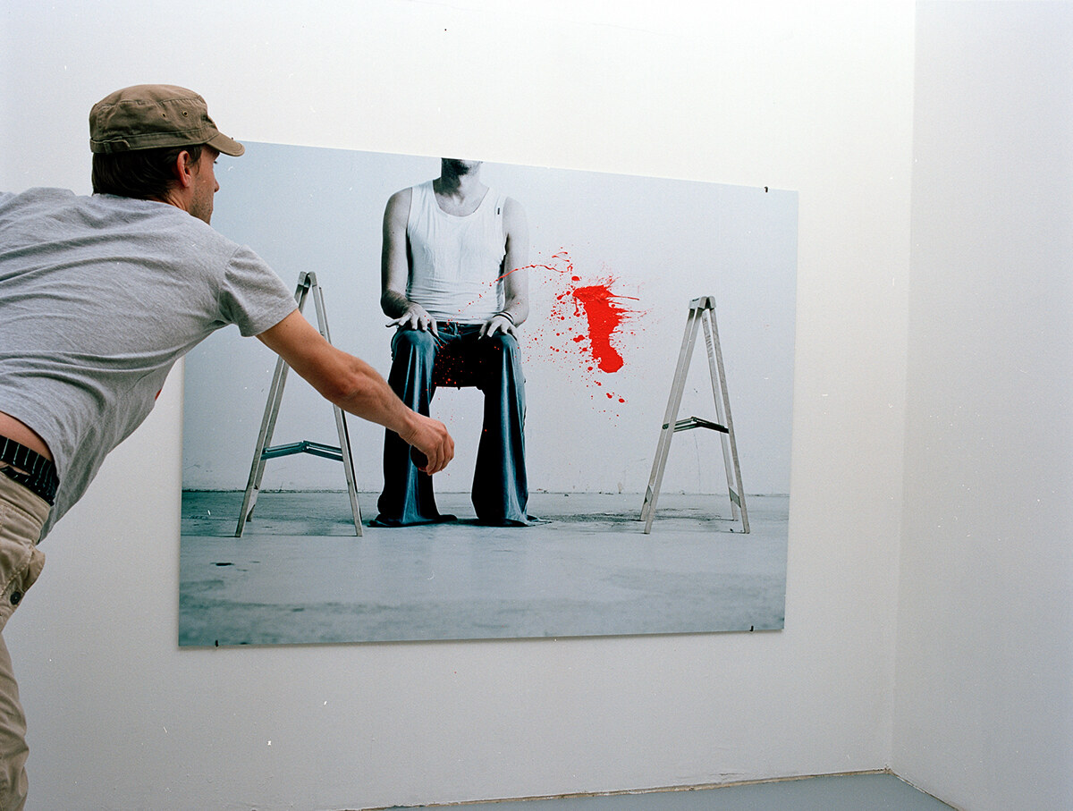 A man in a cap lunging toward the photograph, red paint just thrown across the print