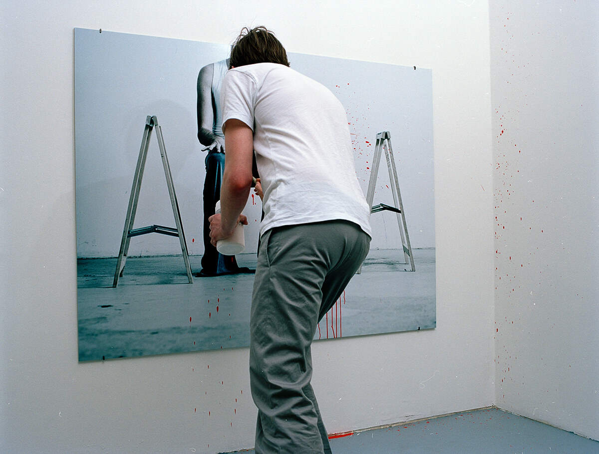 A man crouching low at the photograph with a spray bottle, red drips already running down the print