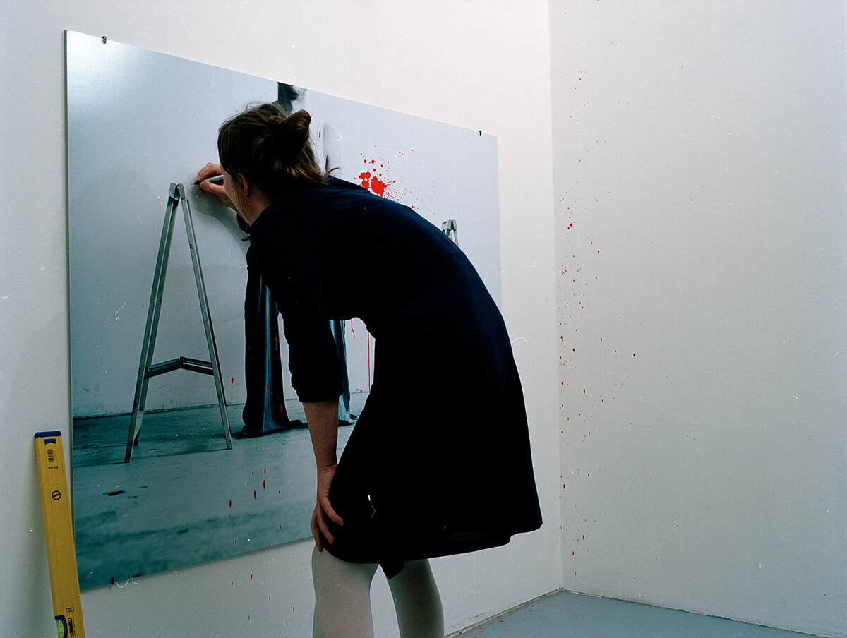 A woman leaning into the top of the photograph, scratching letters into the print, a spirit level resting against the wall