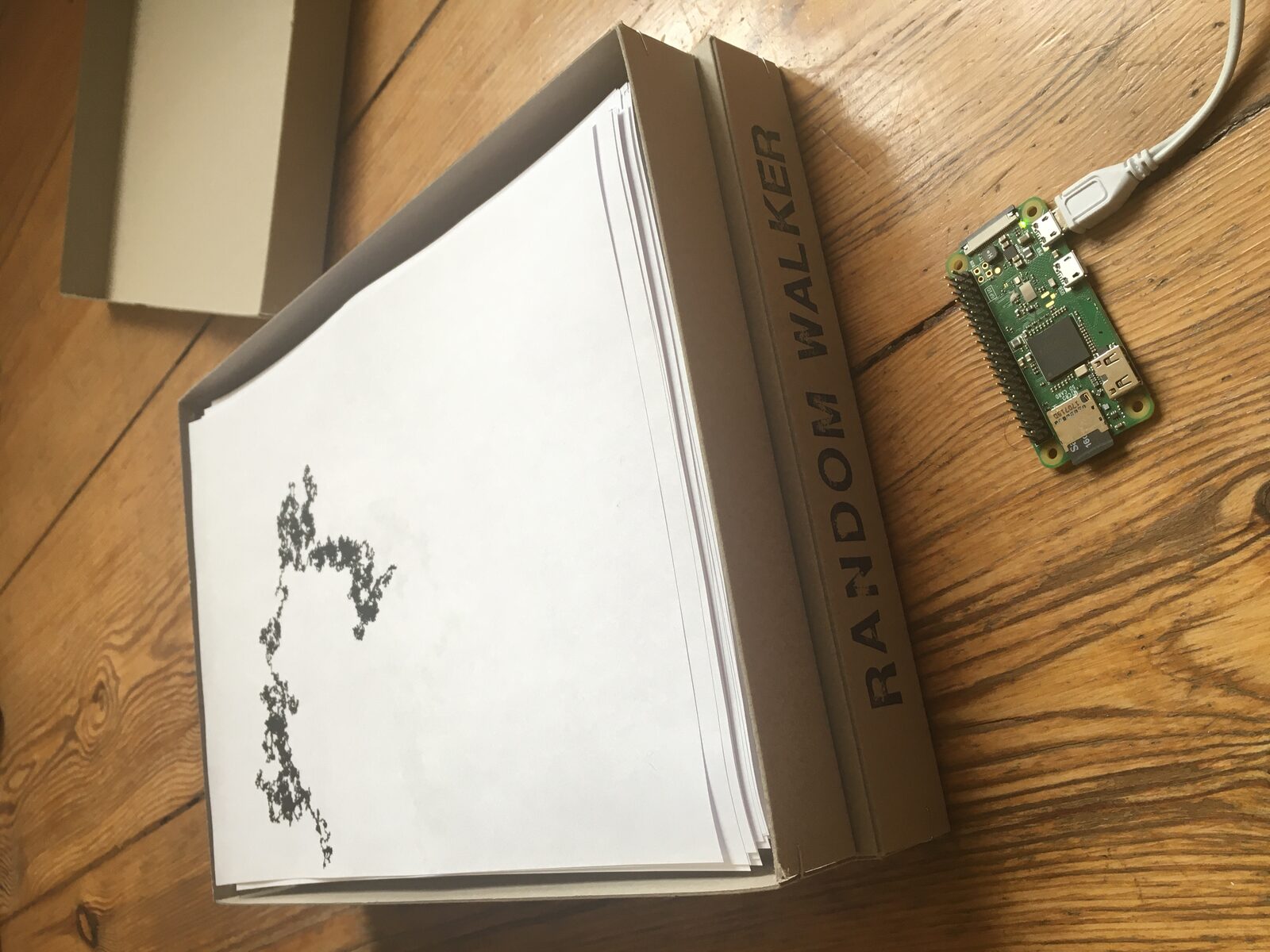 An open archive box spined RANDOM WALKER filled with loose A4 prints, a Raspberry Pi Zero powered beside it