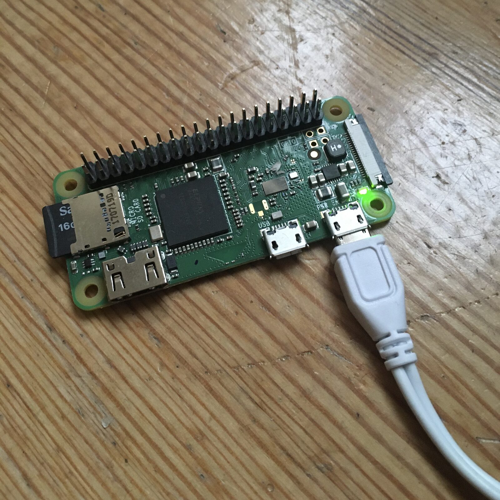 A Raspberry Pi Zero W on a wooden surface, its power LED lit, micro-SD card seated and cable attached