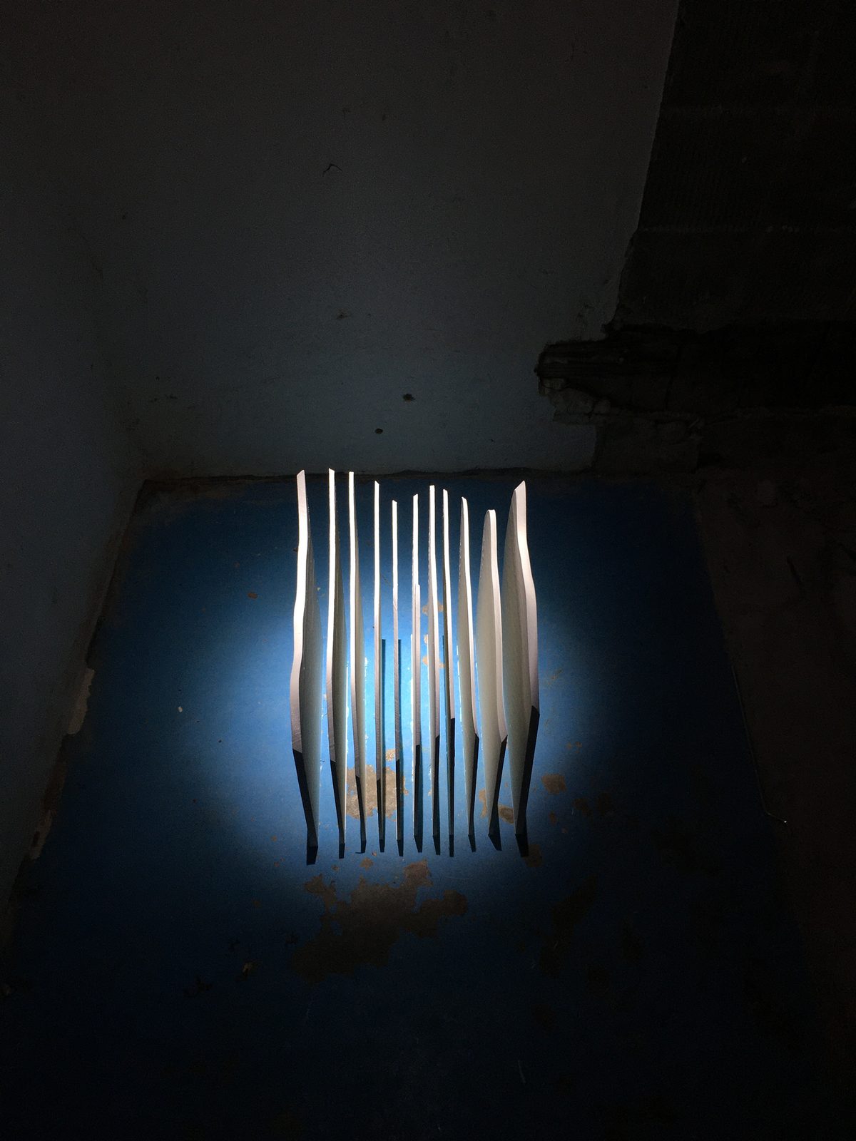 The same room seen wide, the sculpture small in a pool of blue light against a bare corner
